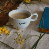 Ceașcă de cafea, o vază cu flori proaspete și o carte deschisă pe o masă luminată de soarele dimineții
