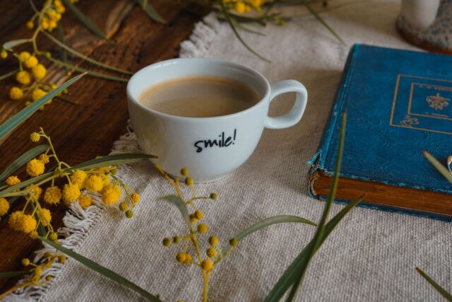 Ceașcă de cafea, o vază cu flori proaspete și o carte deschisă pe o masă luminată de soarele dimineții